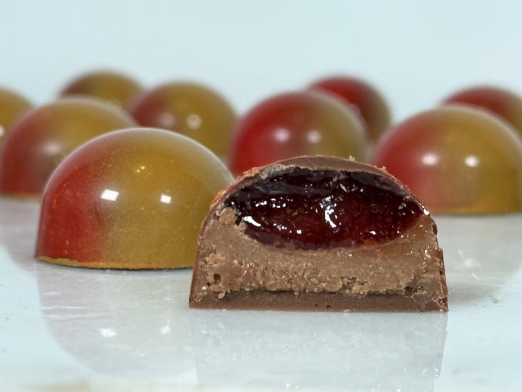A glossy handcrafted chocolate bonbon, bisected to show a layered interior consisting of a smooth, nut paste filling and deep red gel enclosed in a thin milk chocolate shell. Paired with a whole bonbon showing a reflective red and brown exterior.