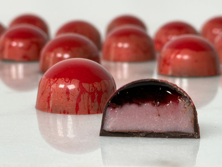 A glossy handcrafted chocolate bonbon, bisected to show a layered interior consisting of a smooth, pink ganache filling and deep red gel enclosed in a thin dark chocolate shell. Paired with a whole bonbon showing a reflective red exterior decorated with red streaks.