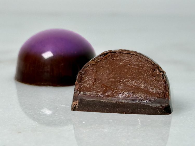 A glossy handcrafted chocolate bonbon, bisected to show a smooth chocolate ganache filling and delicate dark chocolate shell. Paired with a whole bonbon showing a reflective exterior decorated with a purple highlight.