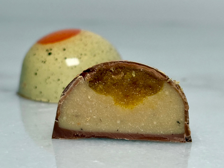 A glossy handcrafted chocolate bonbon, bisected to show a layered interior consisting of a smooth, white chocolate ganache filling and an orange gel enclosed in a thin milk chocolate shell. Paired with a whole bonbon showing a reflective exterior decorated with a central orange circle and black speckles.