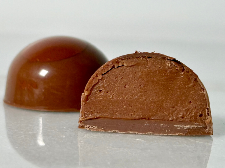 A glossy handcrafted chocolate bonbon, bisected to show a smooth chocolate ganache filling and delicate milk chocolate shell. Paired with a whole bonbon showing a reflective exterior decorated with a light brown highlight.