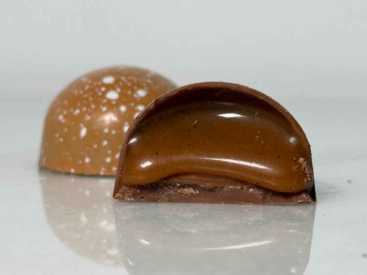 A glossy handcrafted chocolate bonbon, bisected to show a smooth amber caramel filling and delicate milk chocolate shell. Paired with a whole bonbon showing a white speckled decorated exterior.
