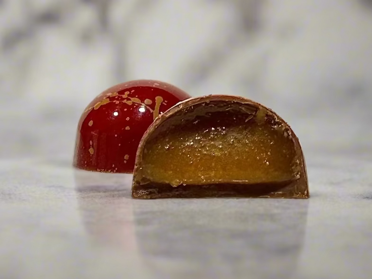 A glossy handcrafted chocolate bonbon, bisected to show a textured amber caramel filling and delicate milk chocolate shell. Paired with a whole bonbon showing a reflective red exterior decorated with golden streaks.