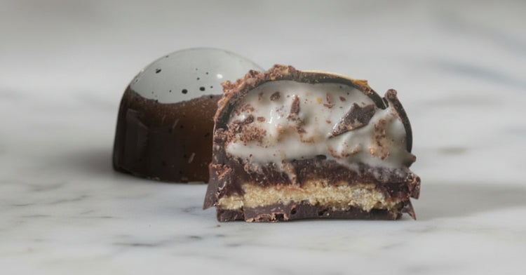 Cross-section of a handcrafted layered chocolate bonbon featuring creamy marshmallow, chocolate ganache, and a delicate cookie layer inside a thin dark chocolate shell. A whole bonbon is to the side, showing a dark brown reflective exterior with a white shiny top.