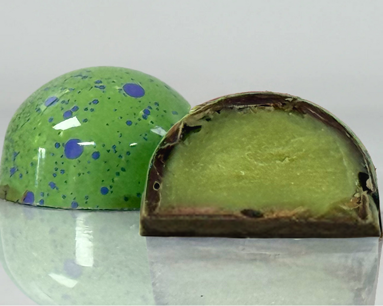 A glossy handcrafted chocolate bonbon, bisected to show a smooth green ganache filling and delicate milk chocolate shell. Paired with a whole bonbon showing a reflective green exterior decorated with blue speckles.