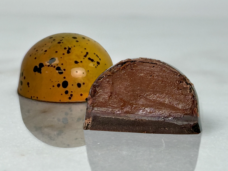 A glossy handcrafted chocolate bonbon, bisected to show a smooth chocolate ganache filling and delicate milk chocolate shell. Paired with a whole bonbon showing a reflective yellow exterior decorated with black speckles.