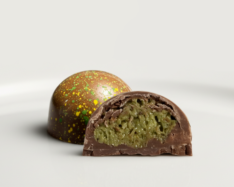 A glossy handcrafted chocolate bonbon, bisected to show a textured green cream filling and delicate milk chocolate shell. Paired with a whole bonbon showing a reflective exterior decorated with green and yellow speckles.