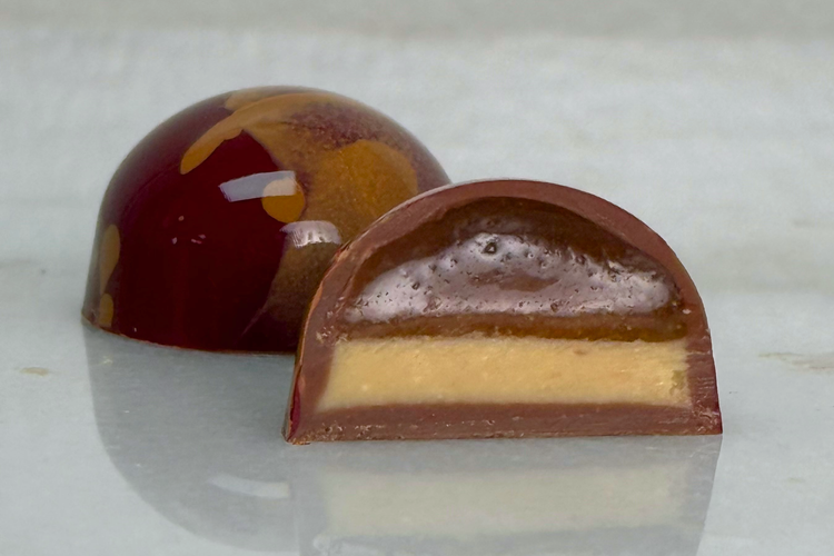 A Peanut Butter Apple Caramel bonbon cut in half beside a whole bonbon, showing its layered filling